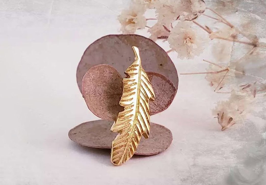 brinco de ouro com formato de pena sobre uma superfície marrom e fundo com flores brancas